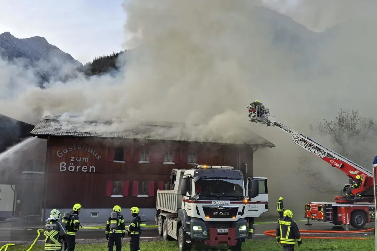 Mehrere Feuerwehrleute sind zu sehen, die einen Brand in einem Holzgebäude bekämpfen. Dichte Rauchwolken steigen auf und ein Feuerwehrfahrzeug ist im Einsatz.