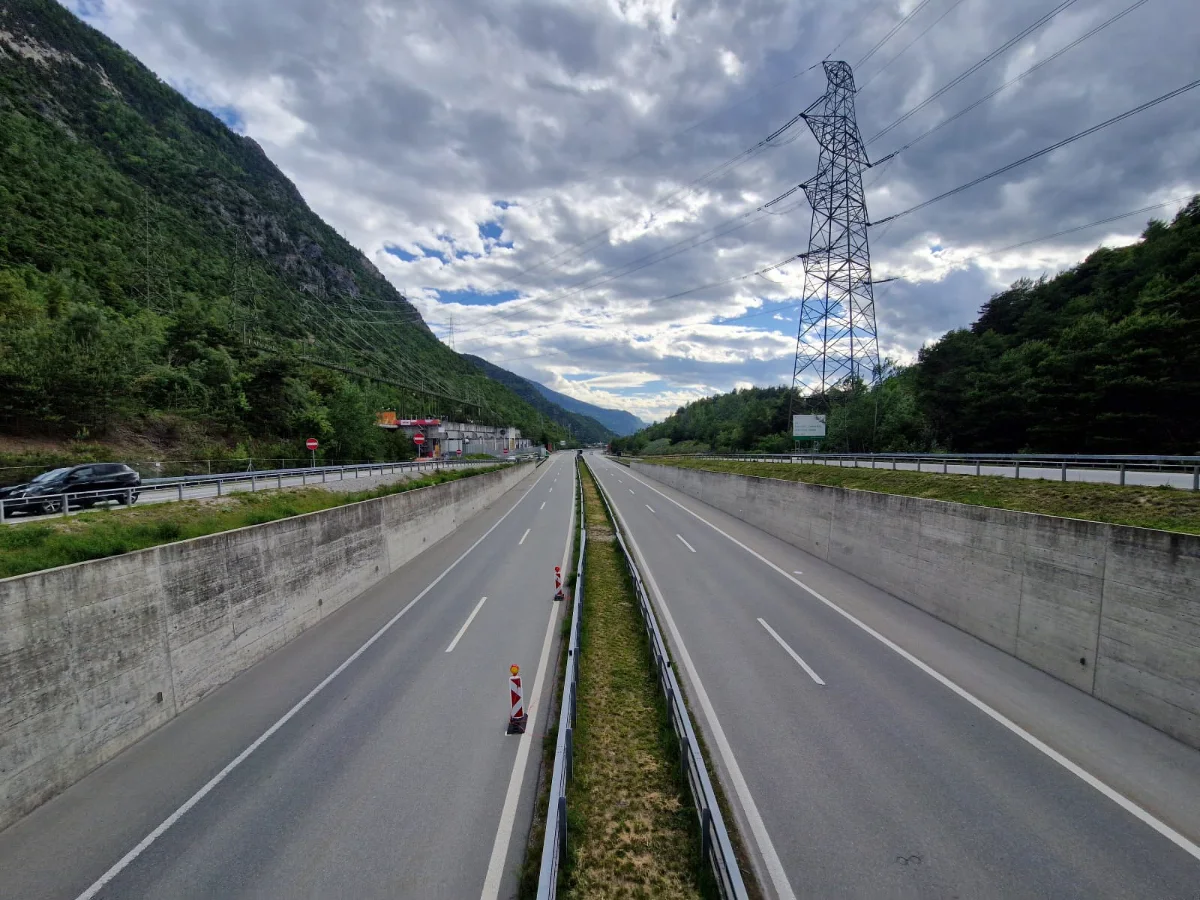 Das Bild zeigt eine verlassene, zweispurige Autobahn in einem bewaldeten Tal, flankiert von Stromleitungen und Bergen. Die Straße ist in gutem Zustand und das Wetter ist bewölkt.