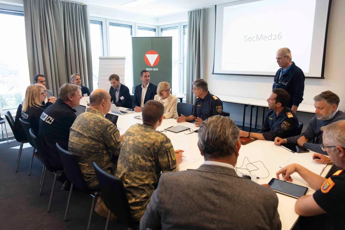 Eine Gruppe von Personen, einige in Uniformen, andere in Zivilkleidung, sitzen um einen Tisch in einem Besprechungsraum. An der Wand hängt ein Bildschirm, auf dem 'SecMed26' steht.