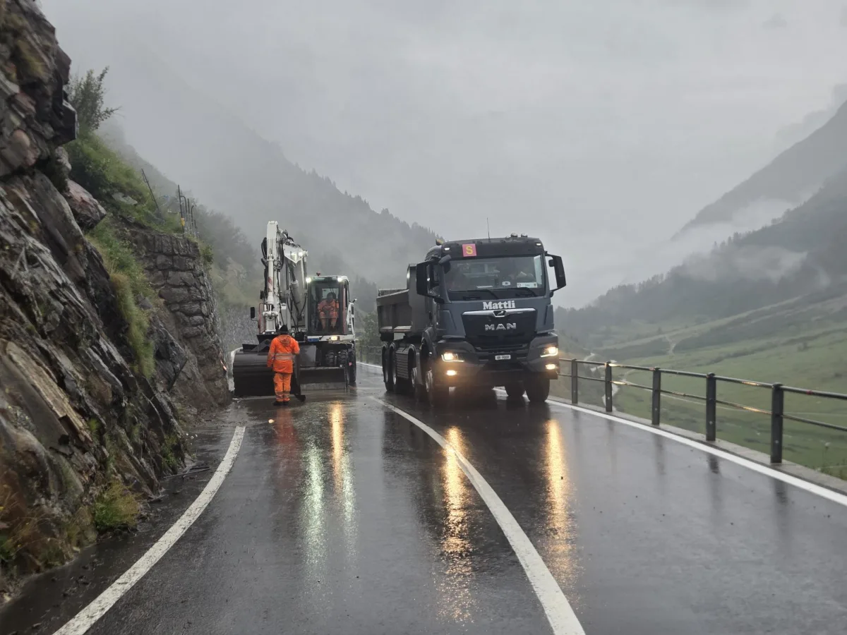 Ein Bagger und ein Lkw stehen auf einer kurvigen Bergstraße. Die Straße ist nass von Regen, während Nebel in den umliegenden Bergen auftritt.