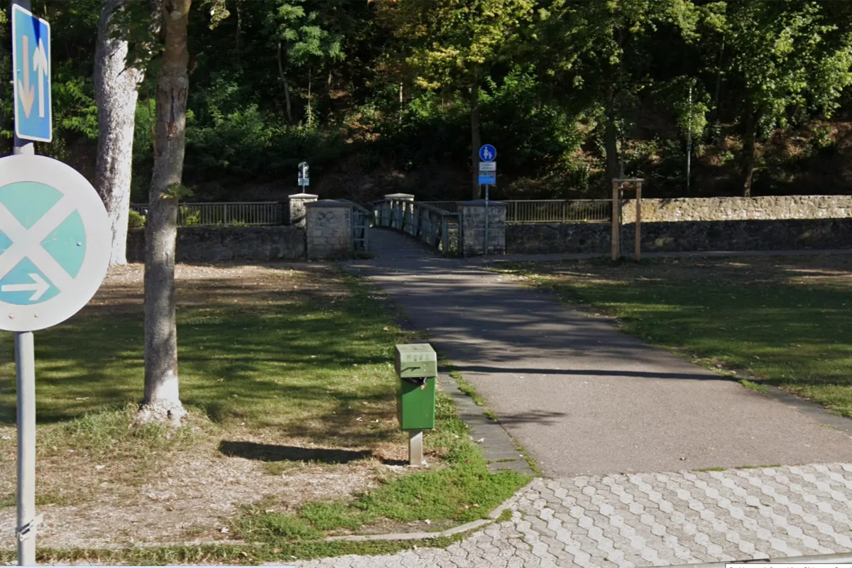 Ein gepflasterter Fußgängerweg führt in Richtung einer steinernen Brücke. Umgeben von Bäumen und Grünflächen strahlt die Umgebung Ruhe aus.