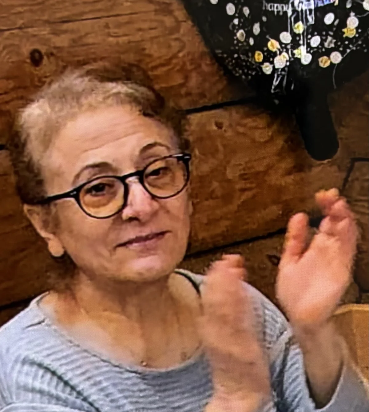 Eine ältere Frau mit Brille sitzt in einem Raum mit Holzwand und klatscht in die Hände. Im Hintergrund ist ein Ballon mit der Aufschrift 'happy' zu sehen.