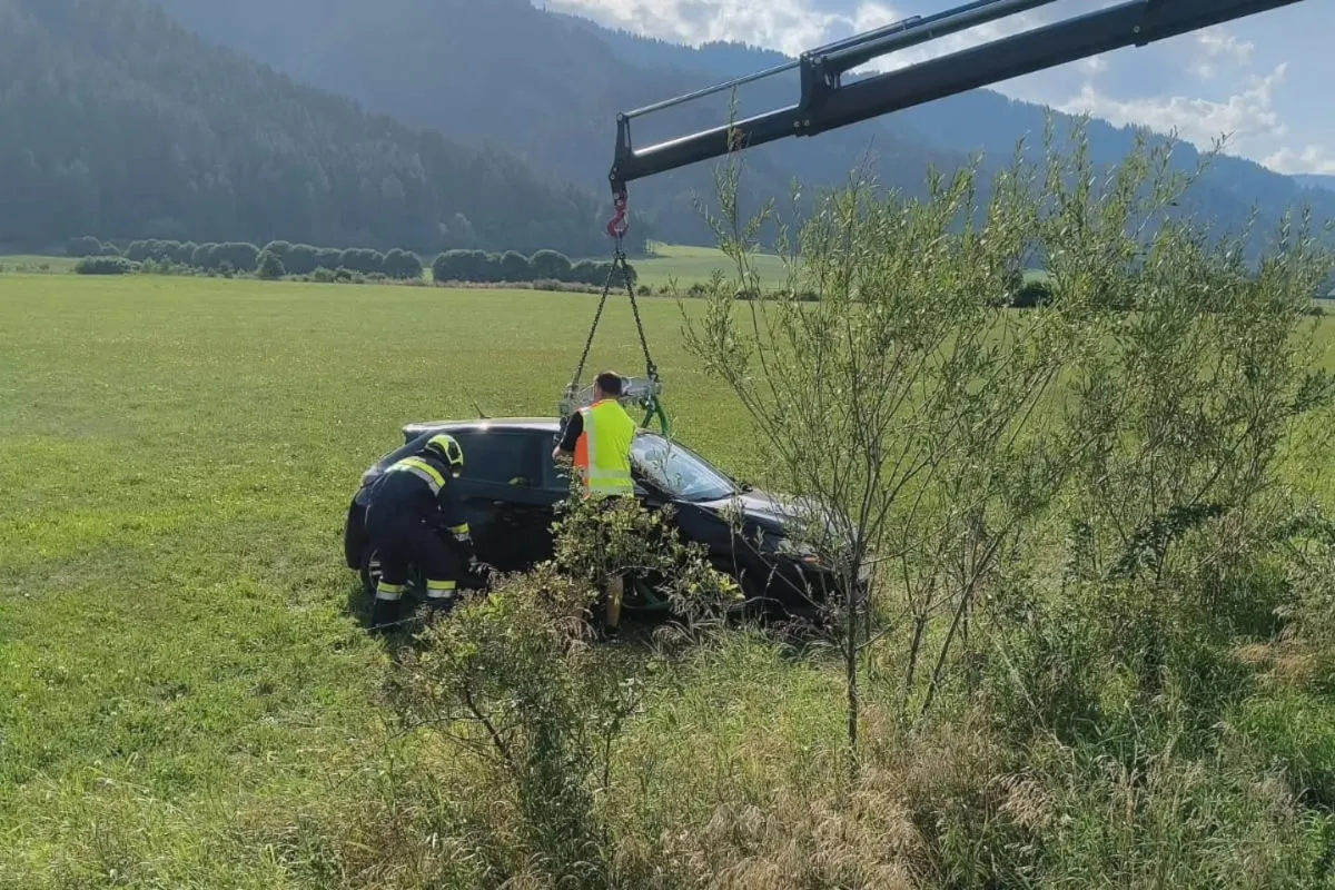 Ein schwarzes Auto wird mit einem Kran aus einem flachen grünen Feld geborgen. Zwei Personen in Warnkleidung sind vor Ort.
