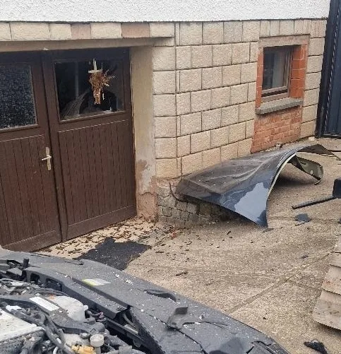 Ein beschädigtes Auto steht vor einer Garage mit einem kaputten Fenster. Autoteile liegen auf dem Boden verstreut.