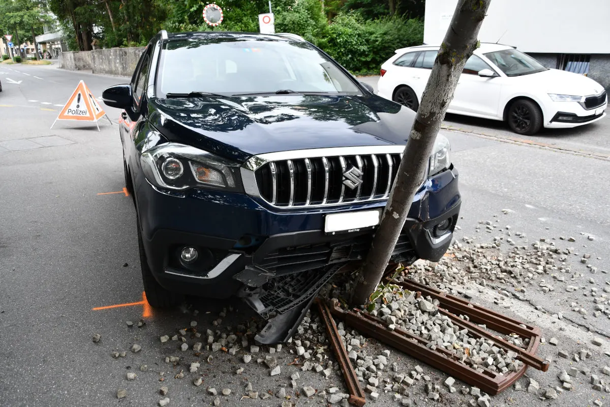 Ein dunkles Auto, vermutlich ein Suzuki, ist frontal gegen einen Baum gefahren. Ein Polizei-Warnschild markiert die Unfallstelle, die in einer städtischen Umgebung liegt.