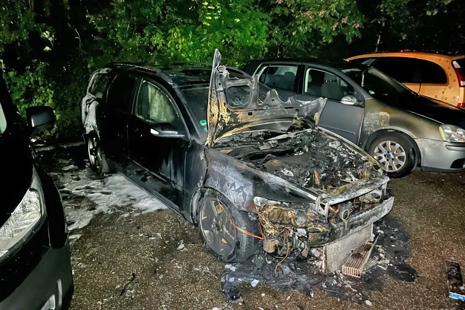Ein stark beschädigtes Auto mit ausgebranntem Motorraum steht auf einem Parkplatz. Weitere Fahrzeuge sind im Hintergrund zu sehen.