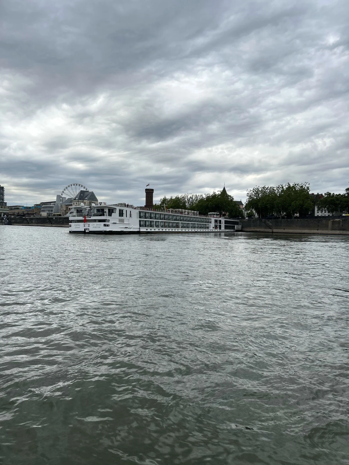 Ein großes weißes Flussschiff fährt auf einem Fluss, im Hintergrund sind städtische Gebäude und ein Riesenrad sichtbar. Der Himmel ist bewölkt.