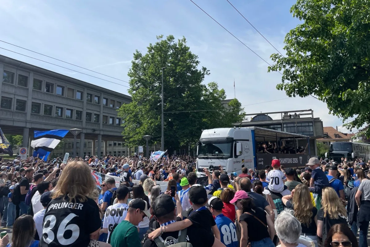 Eine große Menschenmenge steht auf einer Straße und hält Fahnen in der Luft. Ein weißer LKW ist im Hintergrund sichtbar neben einem großen Gebäude.