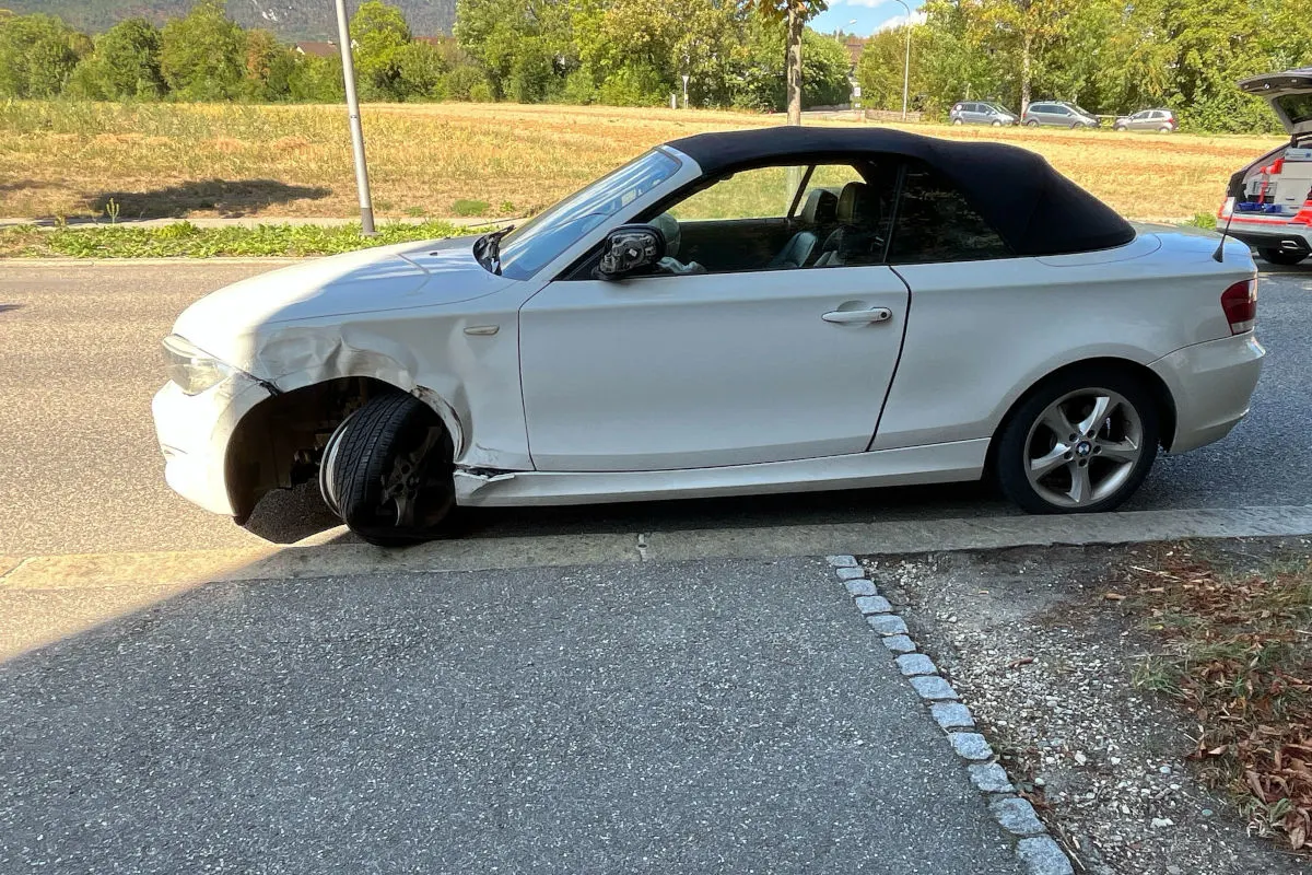 Ein weißes Cabriolet mit einer beschädigten Vorderseite und einem defekten Vorderreifen steht am Straßenrand. Das Auto hat ein schwarzes Stoffverdeck und zeigt eindeutige Kollisionsspuren an der Front.