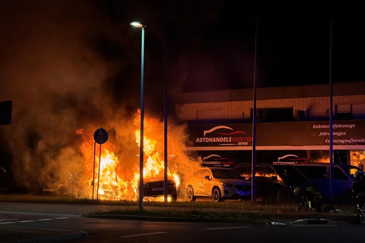 Das Bild zeigt brennende Fahrzeuge vor einem Autohaus bei Nacht. Flammen und Rauch sind gut sichtbar, während ein Teil eines Gebäudes im Hintergrund zu sehen ist.