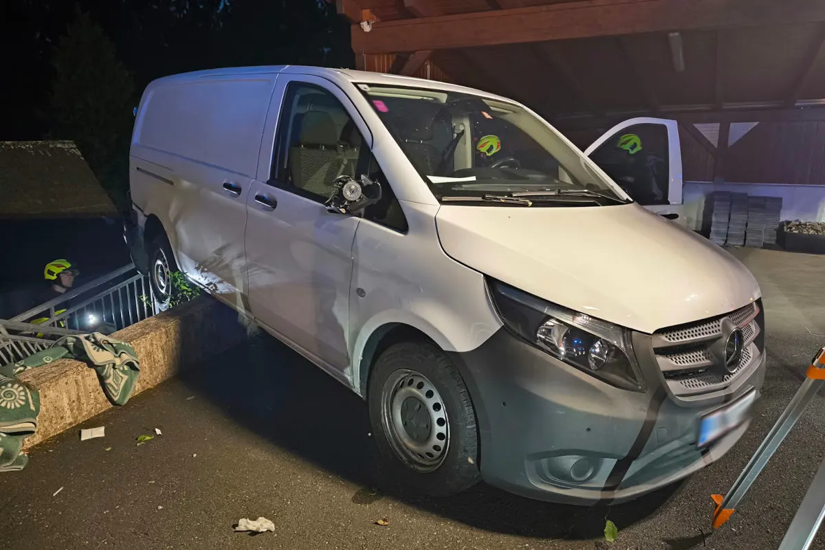 Ein weißer Transporter ist bei Nacht auf einer Betonbarriere auf einem Parkplatz aufgebockt. Ein Feuerwehrmann ist dabei, den Schaden zu begutachten.