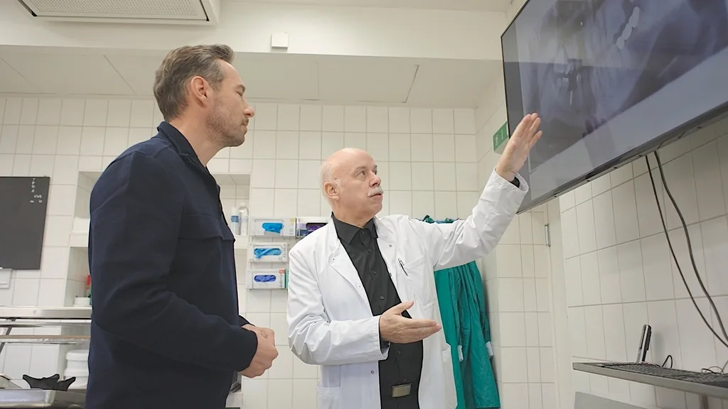 Ein älterer Mann in einem weißen Kittel zeigt auf Röntgenbilder auf einem Bildschirm, während ein anderer Mann daneben steht. Sie befinden sich in einem Labor mit Kacheln und Regalen im Hintergrund.