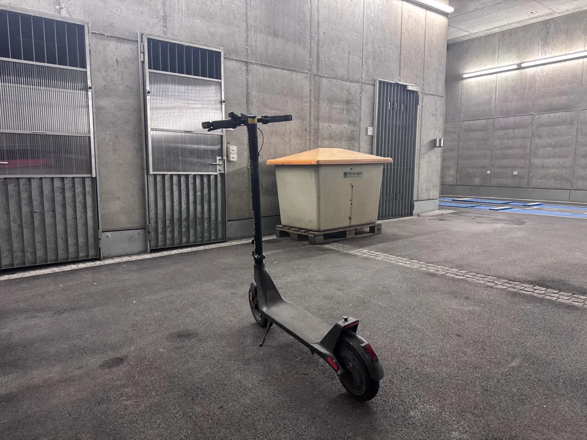 Ein einsamer E-Scooter ist in einem leeren Parkhaus abgestellt. Die Umgebung besteht aus Betonwänden und einem großen Abfallcontainer.