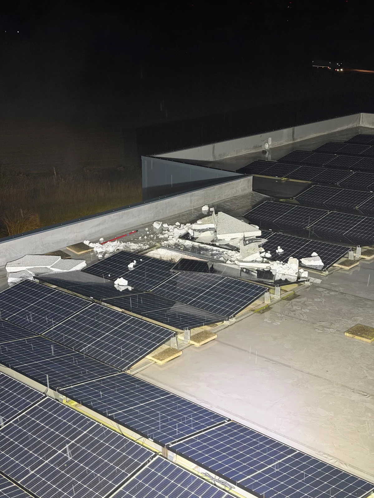 Zerstörte Solarmodule und Trümmerteile sind auf einem Dach zu sehen. Es ist Nacht, und im Hintergrund ist die Straße schwach erleuchtet.
