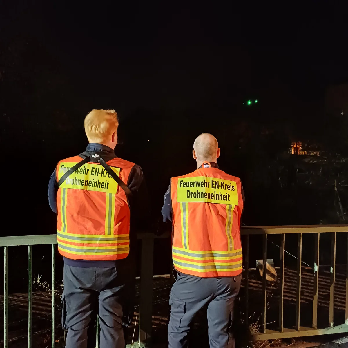Zwei Personen in orangefarbenen Westen mit der Aufschrift 'Feuerwehr EN-Kreis Drohneneinheit' stehen nachts an einem Geländer. Der Hintergrund ist dunkel.