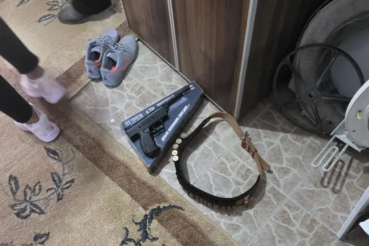 Auf dem Boden neben einem Paar Schuhe liegt eine Verpackung mit einer Spielzeugpistole und ein Gürtel mit Patronenattrappen.
