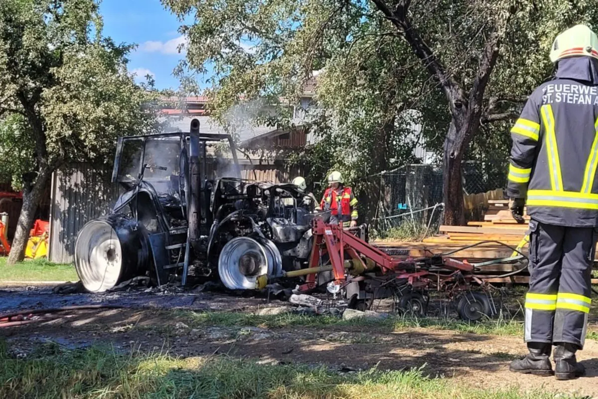Ein verbrannter Traktor steht auf einer Wiese, umgeben von zwei Feuerwehrleuten in Schutzkleidung.