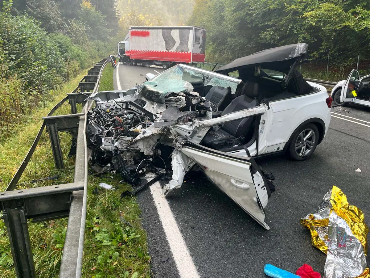 Ein stark beschädigtes weißes Cabriolet steckt in einer Leitplanke auf einer Landstraße. Ein LKW steht im Hintergrund.