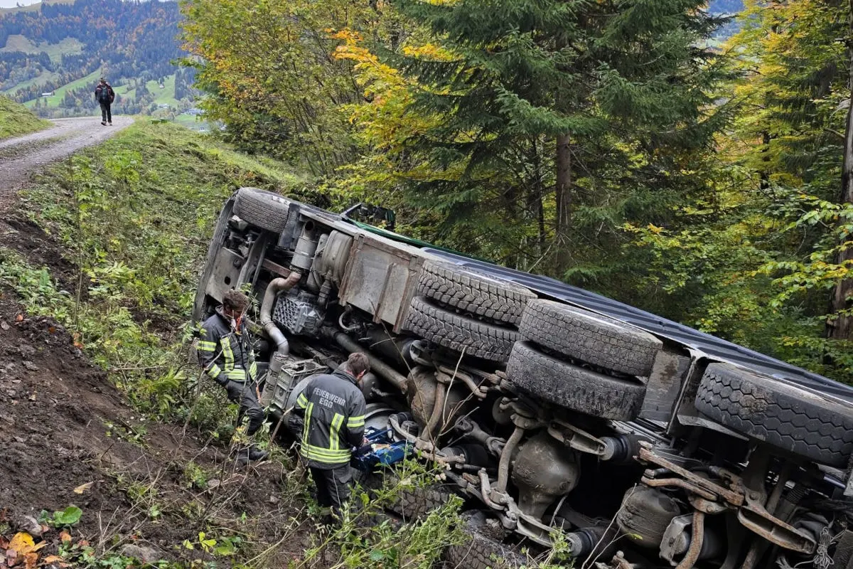 Ein großer Lastwagen liegt auf der Seite neben einer Böschung. Zwei Feuerwehrleute in Einsatzkleidung überprüfen die Situation.