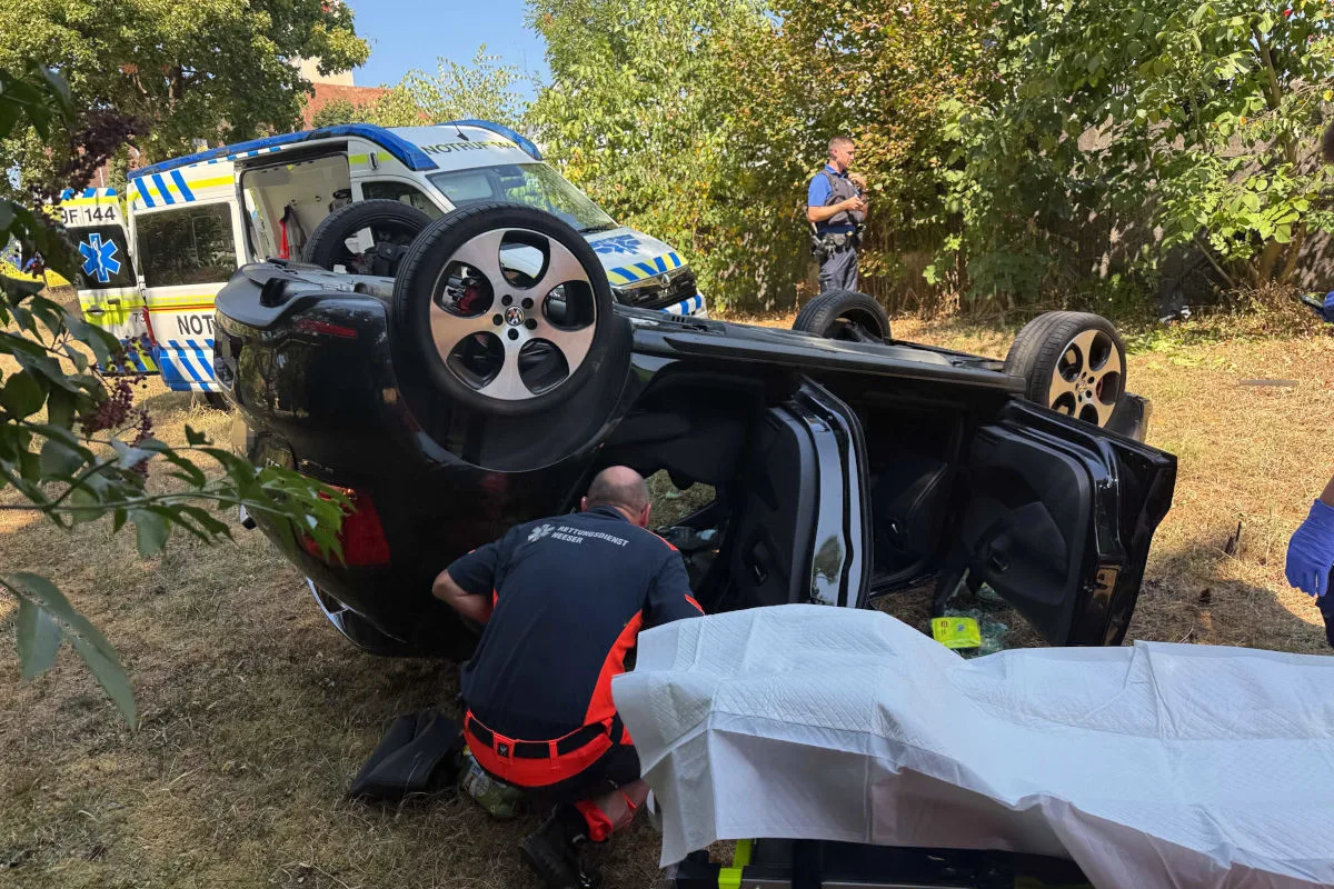 Ein Auto ist auf dem Dach in einem grasigen Bereich mit Rettungskräften drumherum. Ein Notarztwagen mit offener Tür ist im Hintergrund erkennbar.