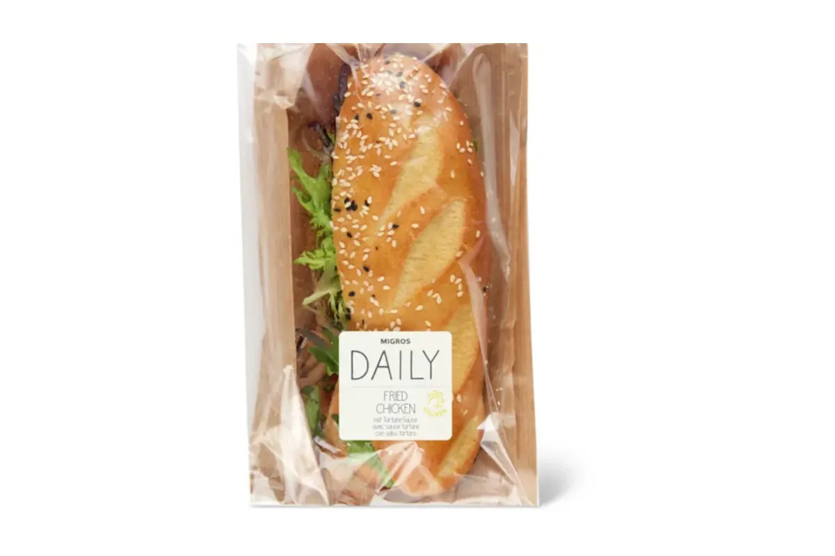 Ein Sandwich mit Sesambrötchen ist in einer Klarsichtfolie verpackt. Auf der Verpackung steht 'Migros Daily'.