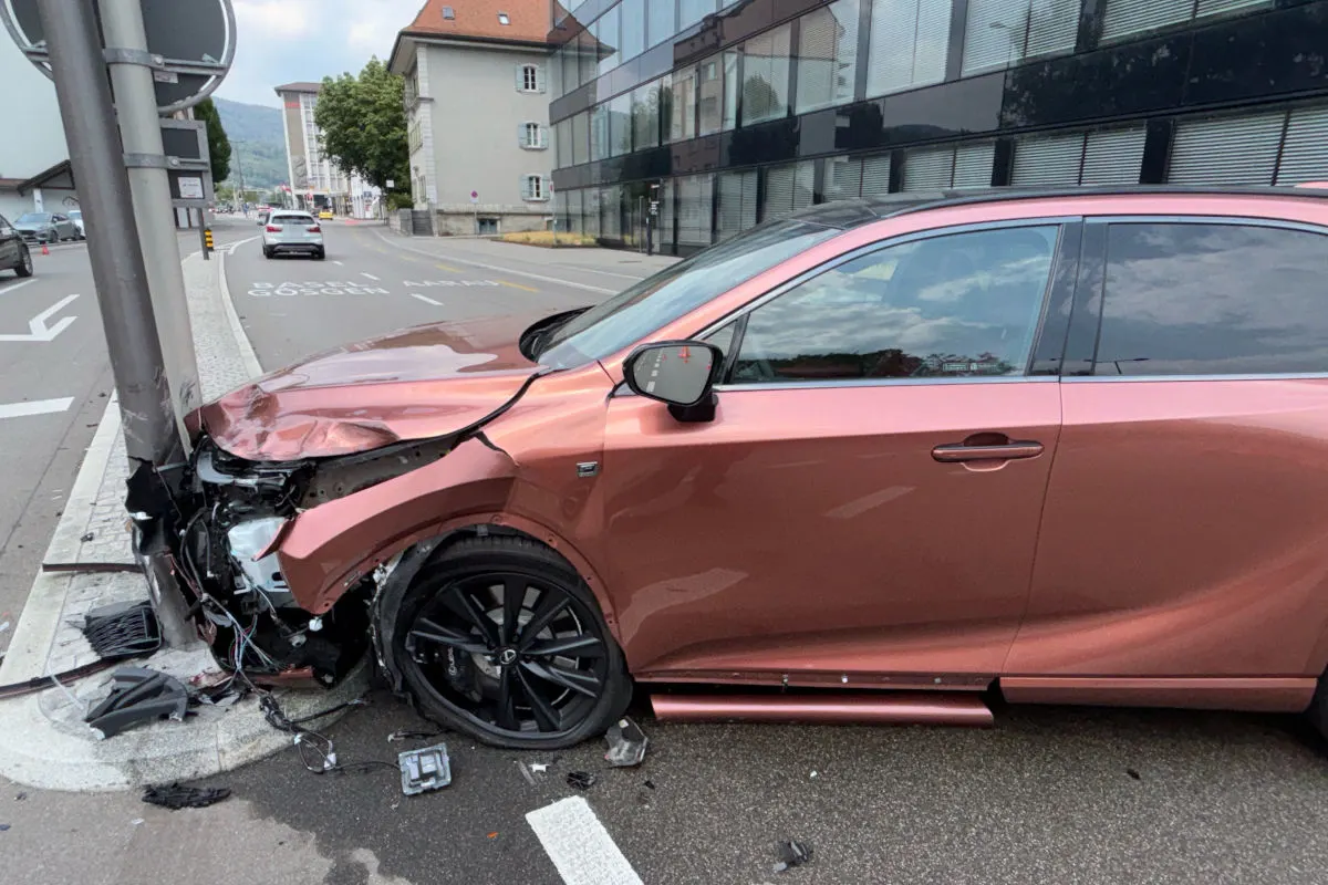 Ein rosafarbener Pkw ist frontal beschädigt, nachdem er gegen einen Mast am Straßenrand gefahren ist. Der Mast zeigt deutliche Spuren des Aufpralls am Fahrzeug.