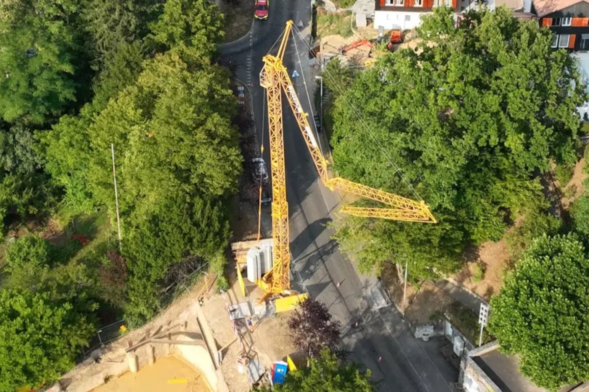 Ein gelber Baukran ist umgeknickt und liegt quer über eine Straße in einem von Bäumen umgebenen Wohngebiet. Mehrere Fahrzeuge sind in der Nähe des Kranes zu sehen.