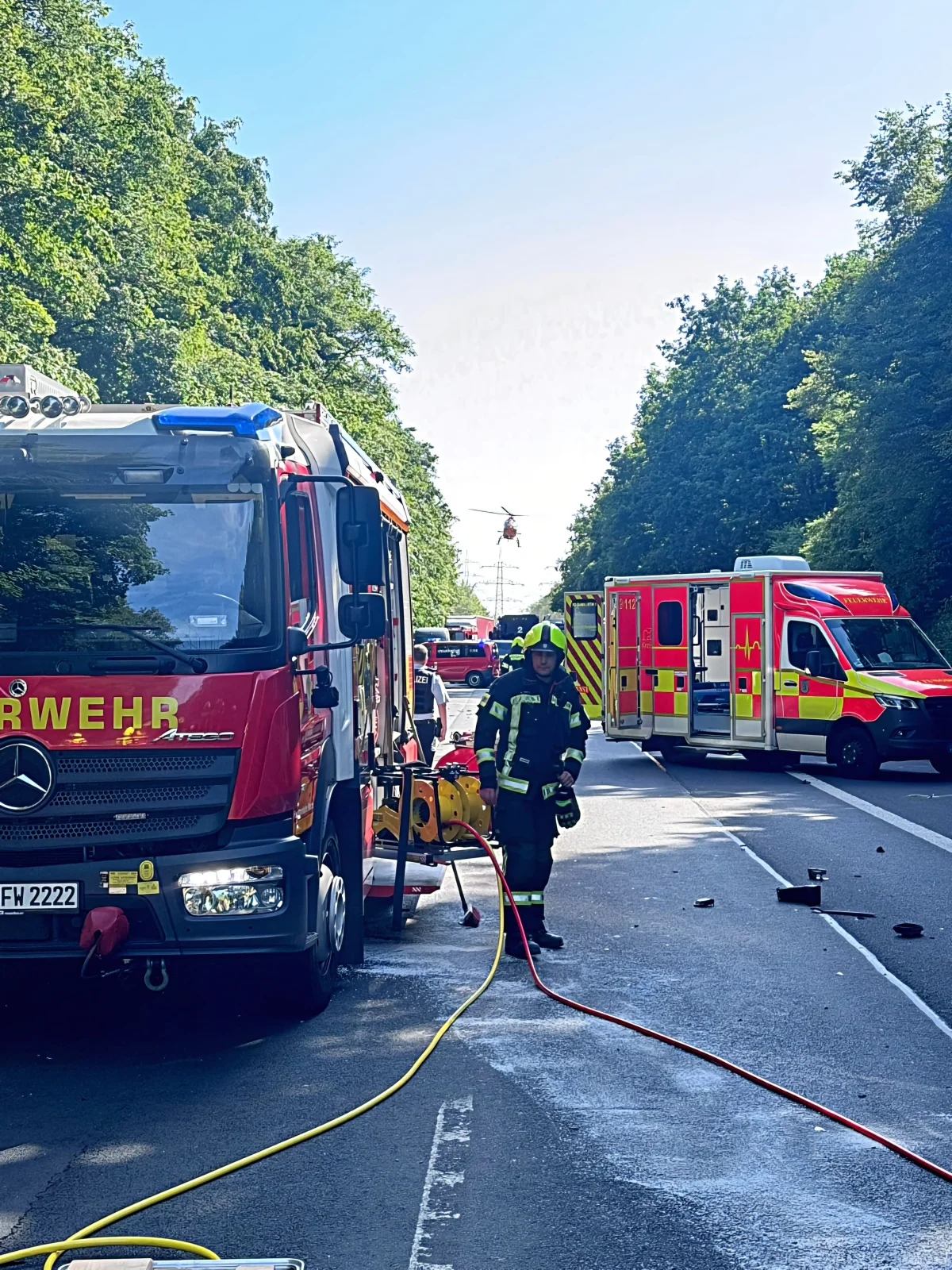 Ein Feuerwehrfahrzeug und mehrere Rettungswagen stehen auf einer von Bäumen gesäumten Straße, ein Feuerwehrmann bewegt sich dazwischen.