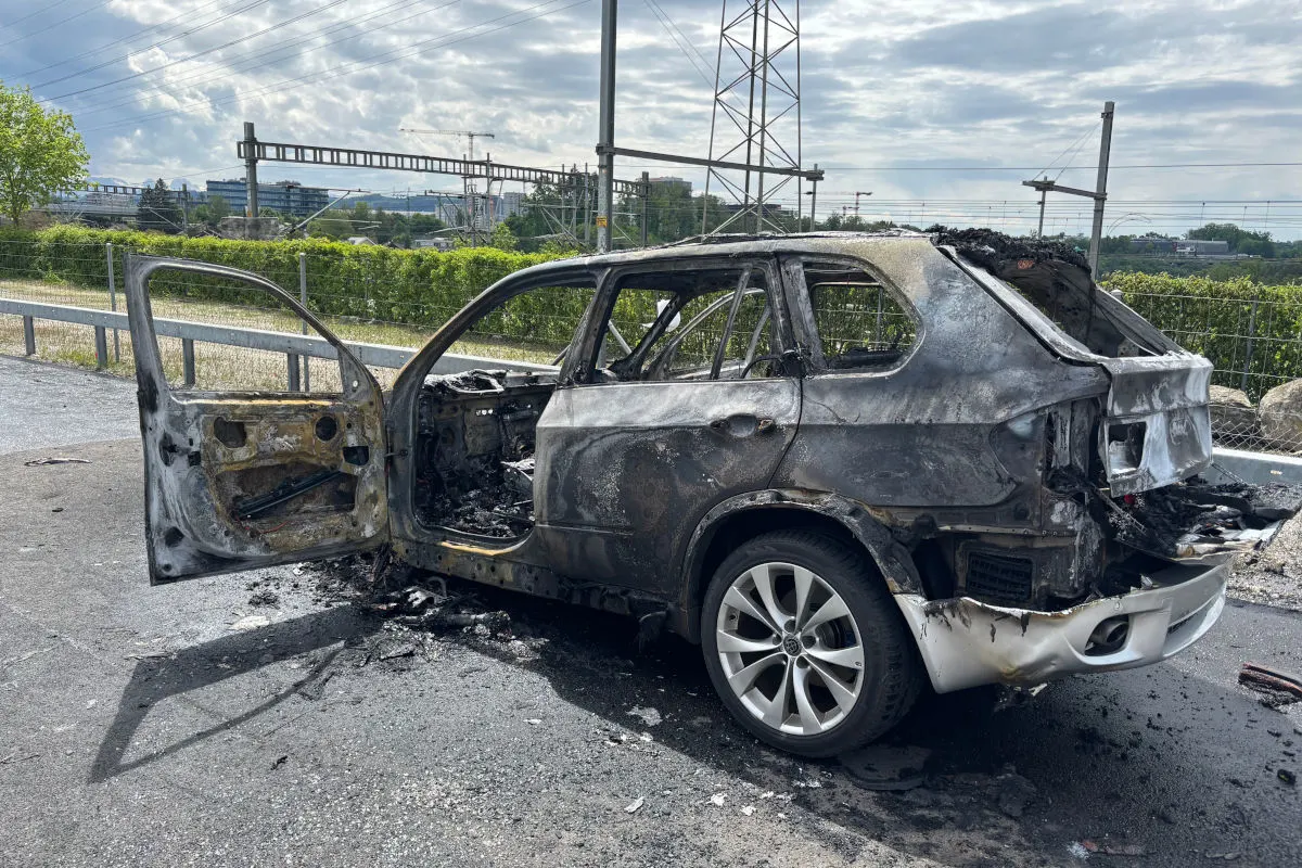 Ein vollständig ausgebranntes Auto steht mit geöffneter Tür auf einer Straße. Im Hintergrund sind Bahngleise und Infrastruktur zu sehen.