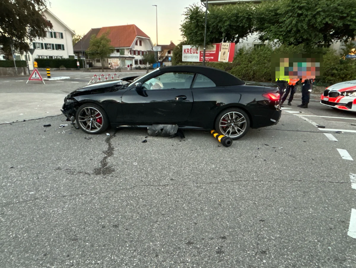 Ein schwarzes Auto mit erheblichen Frontschäden steht auf einer Straße. Polizeibeamte sind rechts im Hintergrund erkennbar.