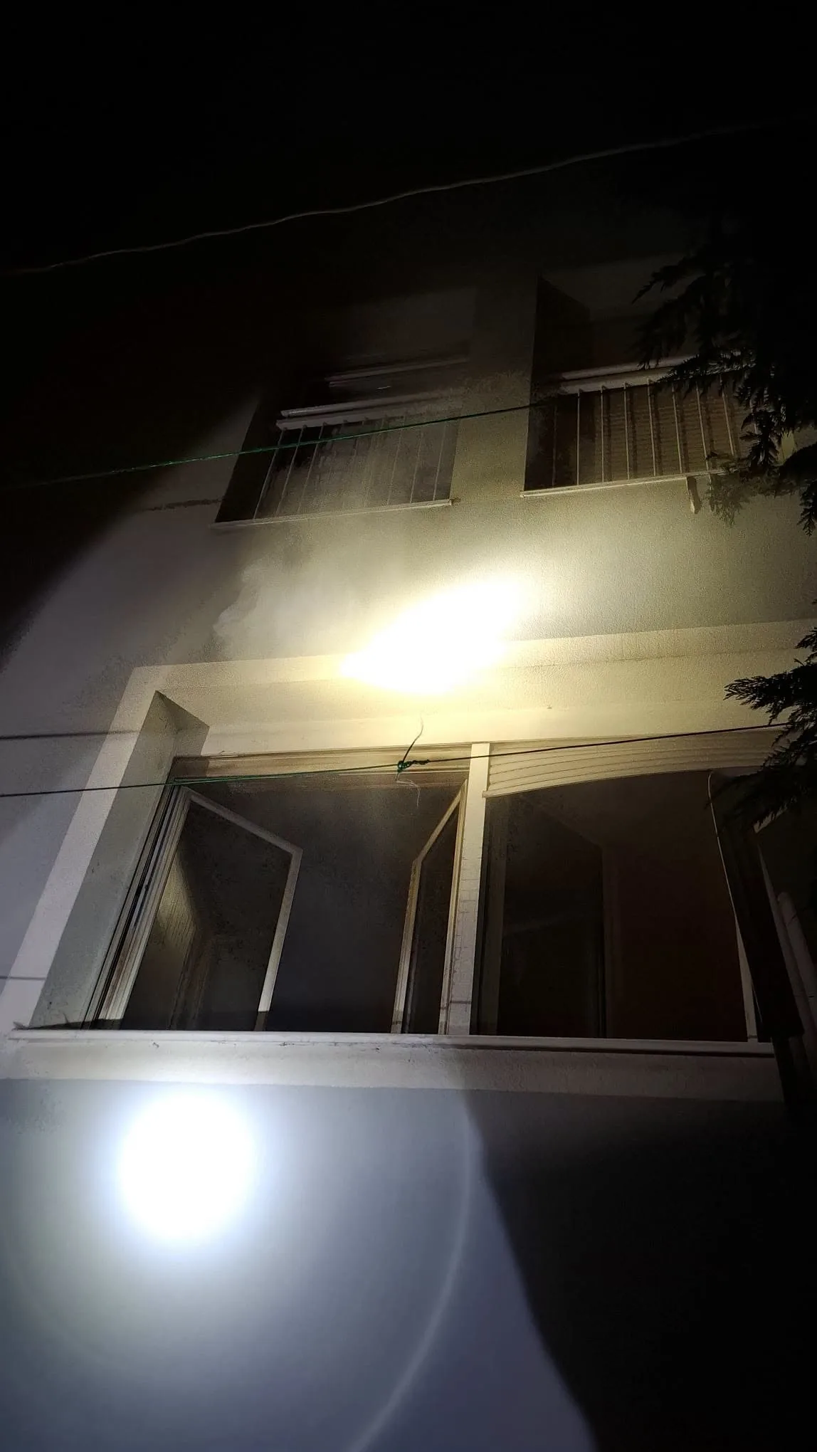 Ein mehrstöckiges Wohnhaus wird bei Nacht beleuchtet, während ein offenes Fenster sichtbar ist. Das Licht stammt möglicherweise von einer Taschenlampe oder einem Einsatzfahrzeug.