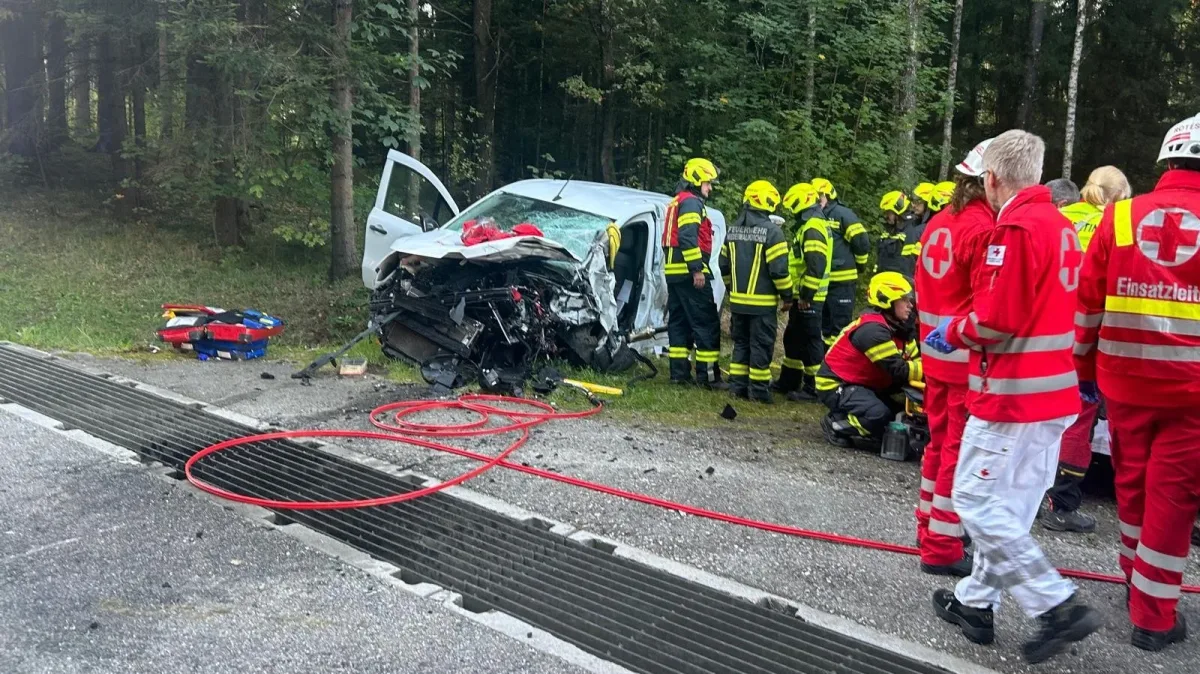 Ein stark beschädigtes Auto steht am Straßenrand eines Waldgebiets. Mehrere Rettungskräfte in Schutzkleidung sind darum versammelt.