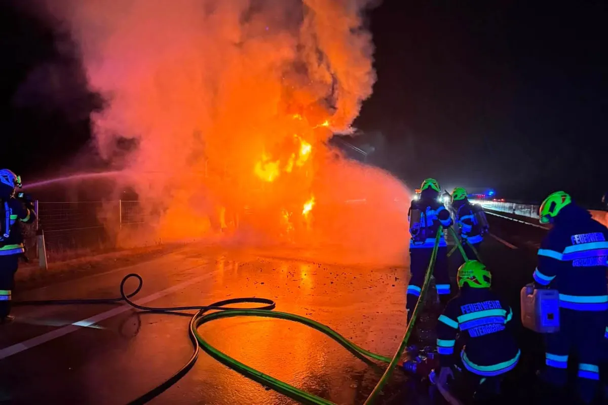 Mehrere Feuerwehrleute löschen einen großen Fahrzeugbrand auf einer Autobahn. Es ist Nacht, und der Brand erzeugt große orangefarbene Flammen und Rauch.