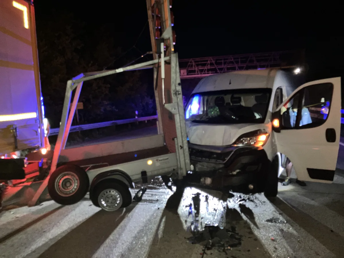 Ein weißer Lieferwagen ist frontal mit einem Anhänger auf einer Autobahn kollidiert. Die Fahrertür ist geöffnet, es ist Nacht.