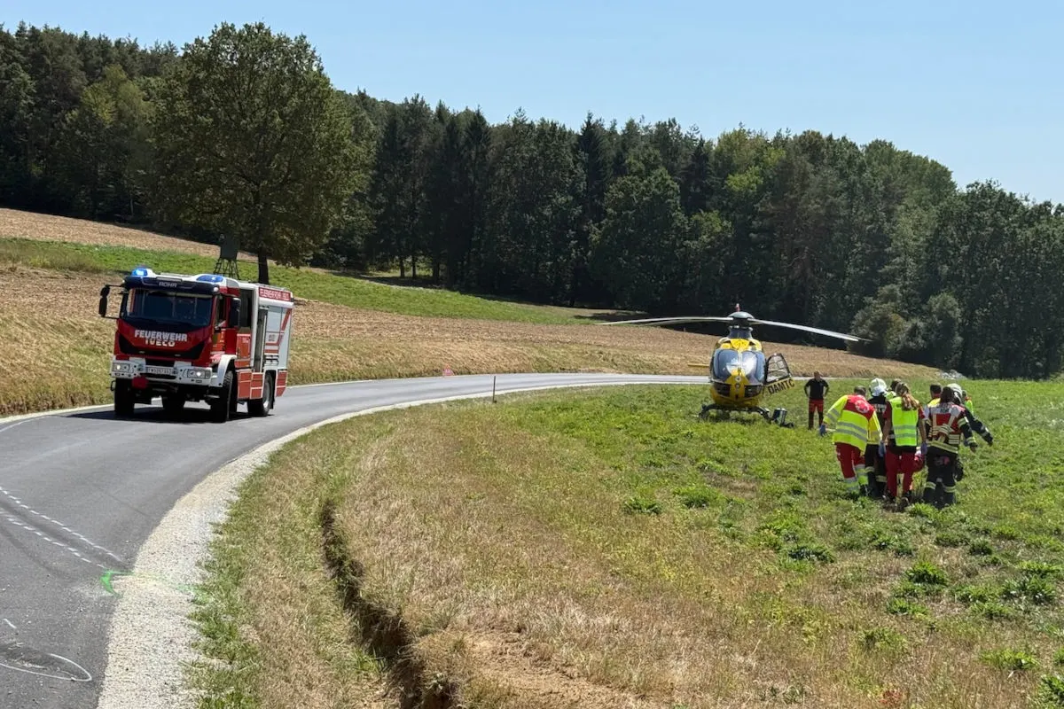 Ein rotes Feuerwehrauto fährt auf einer asphaltierten Straße, während ein gelber Rettungshubschrauber auf einem angrenzenden Feld gelandet ist. Rettungskräfte in leuchtender Kleidung begeben sich zum Hubschrauber.