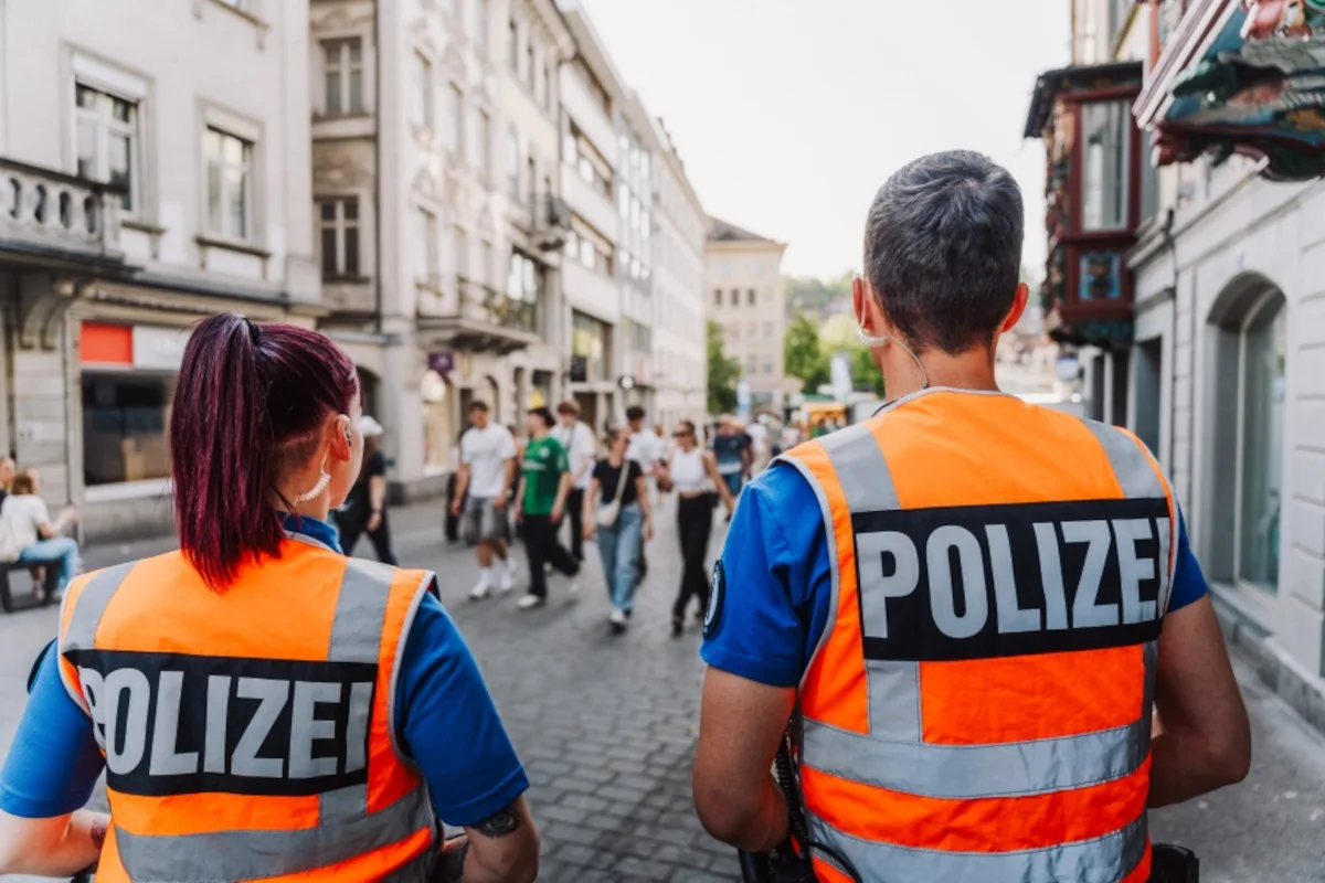 Zwei Polizisten in orangefarbenen Warnwesten stehen in einer belebten Fußgängerzone. Mehrere Menschen gehen im Hintergrund.