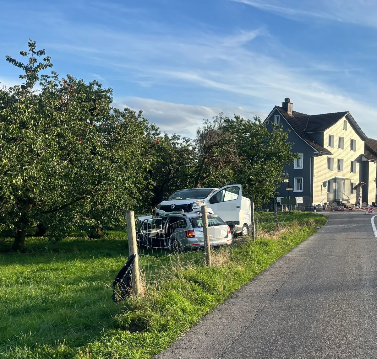 Ein weißer Transporter steht auf einem geparkten silbernen Pkw neben einer Landstraße. Im Hintergrund sind Bäume und ein Wohnhaus zu sehen.