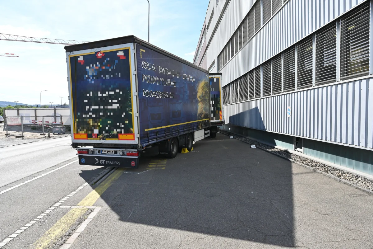 Ein Lastwagenanhänger steht dicht an einem großen Gebäude auf einem Bürgersteig. Der Anhänger scheint gegen die Wand gedrückt zu sein.