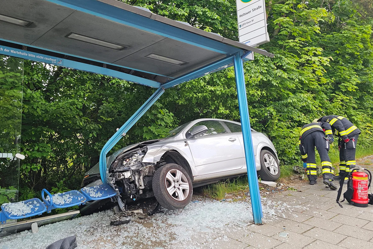Ein silbernes Auto ist in eine Bushaltestelle gefahren, mit Glassplittern am Boden. Zwei Einsatzkräfte arbeiten an der Unfallstelle.
