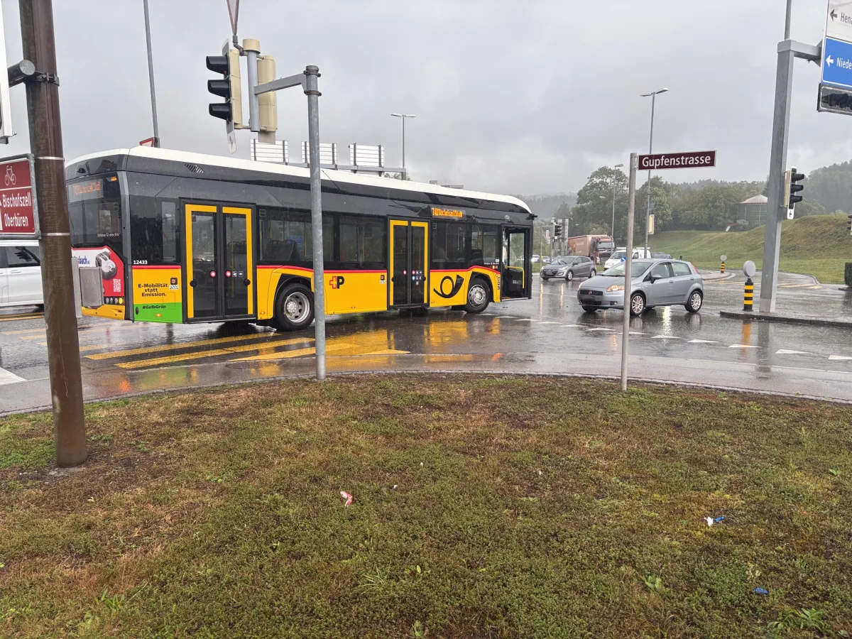Ein gelb-schwarzer Linienbus steht bei Regen an einer Kreuzung. In der Nähe befinden sich mehrere Autos und Verkehrszeichen.