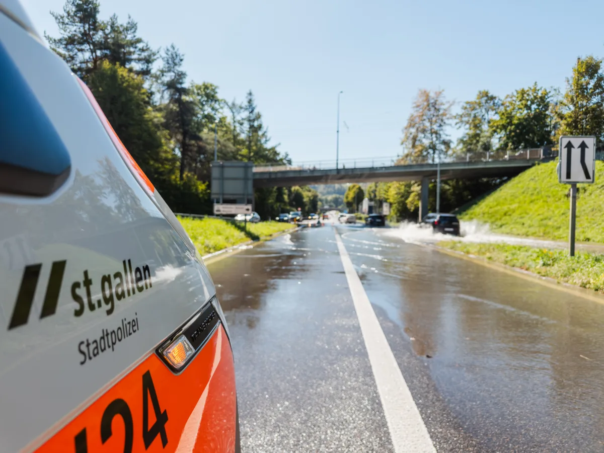Auf einer nassen Straße in St. Gallen steht ein Polizeifahrzeug der Stadtpolizei nahe einer Brücke. Autos spritzen Wasser.