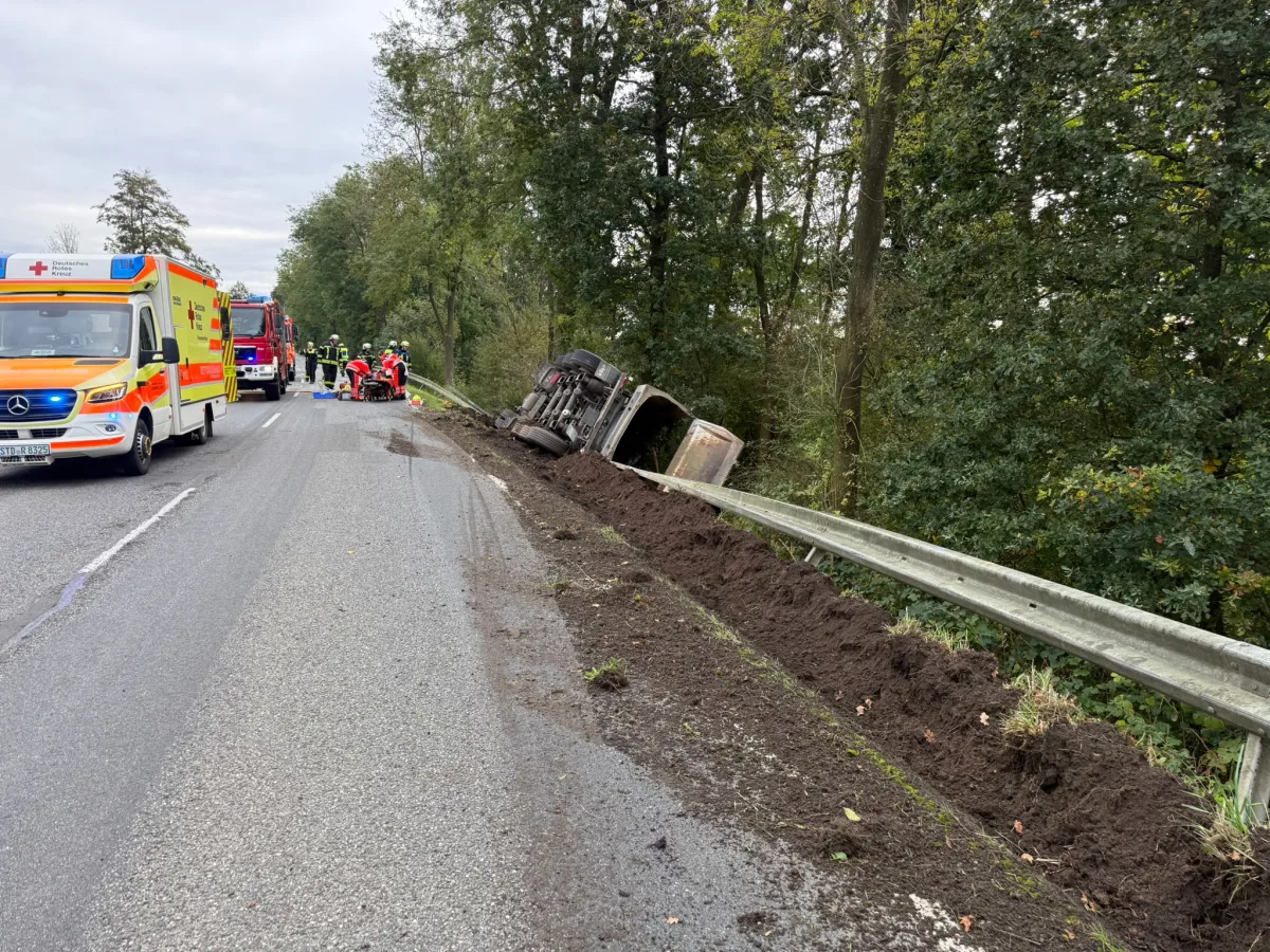 Ein umgekippter Lkw liegt neben der Straße, während Rettungskräfte bei einem Krankenwagen tätig sind. Im Hintergrund sind Bäume sichtbar.