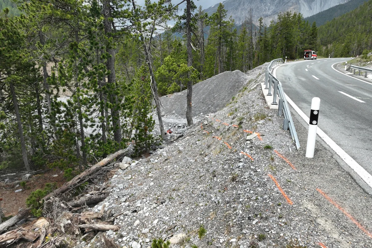 Eine Bergstraße führt an einem Waldstück vorbei, wo ein Fahrzeug am Straßenrand steht. Die Umgebung ist durch eine leichte Schräge und vereinzelte Bäume gekennzeichnet.