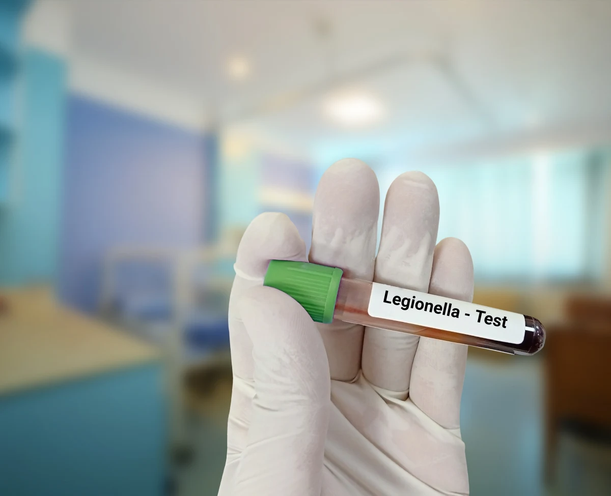 In einem Labor hält eine behandschuhte Hand ein Röhrchen mit der Aufschrift 'Legionella - Test'. Im Hintergrund sind Laborgeräte unscharf zu sehen.
