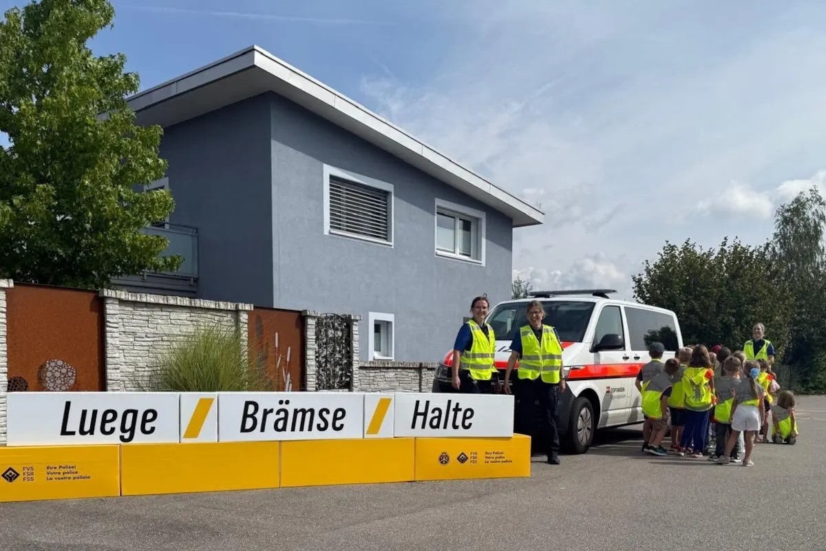 Zwei Polizisten in Warnwesten stehen mit einer Gruppe von Kindern neben einem Polizei-Van vor einem modernen Haus.
