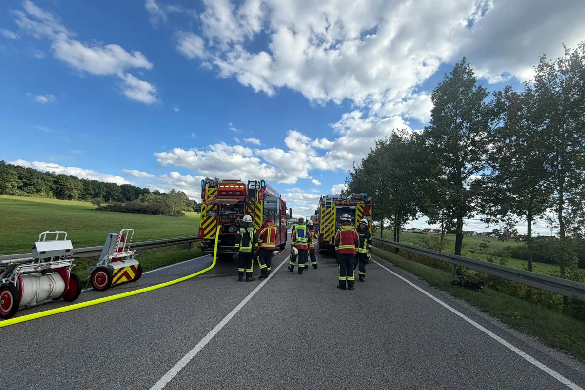 Zwei Feuerwehrfahrzeuge stehen mit mehreren Einsatzkräften auf einer Landstraße. Der Himmel ist blau mit wenigen Wolken.