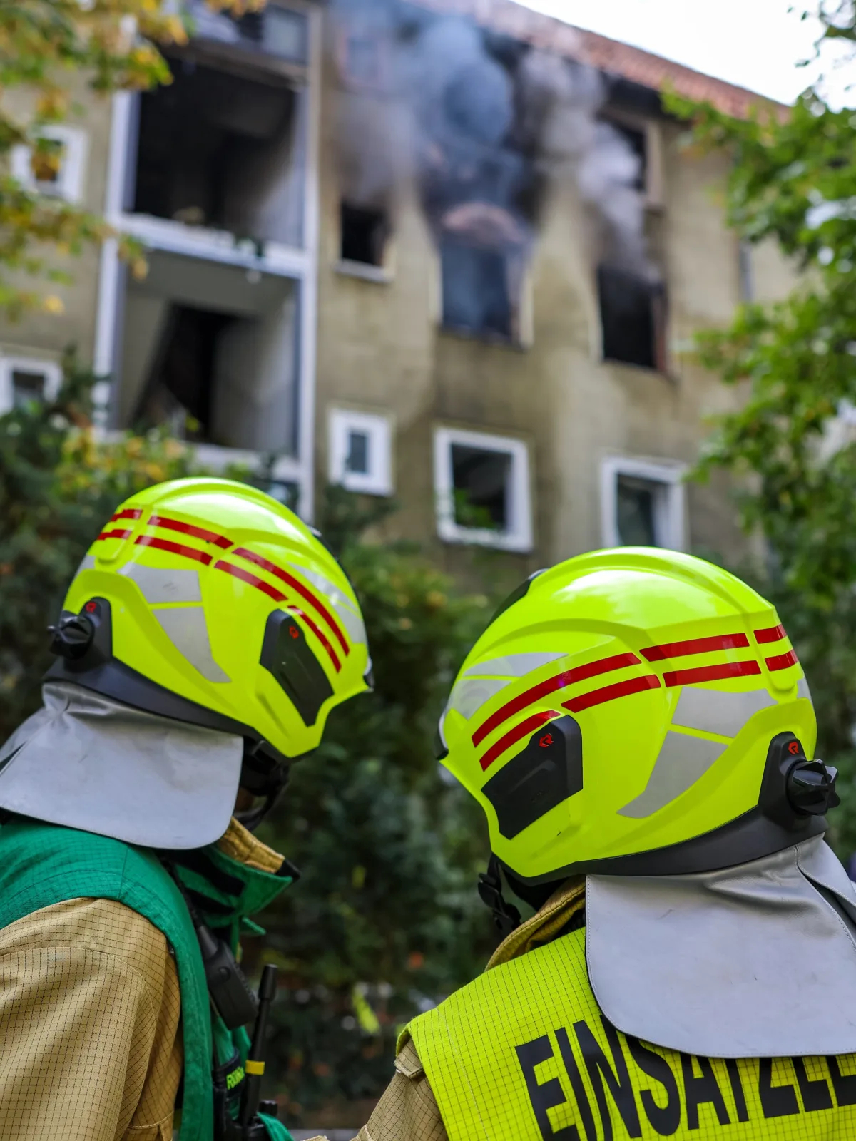 Zwei Feuerwehrleute in gelben Helmen stehen vor einem brennenden Mehrfamilienhaus. Aus den Fenstern quillt dichter Rauch.