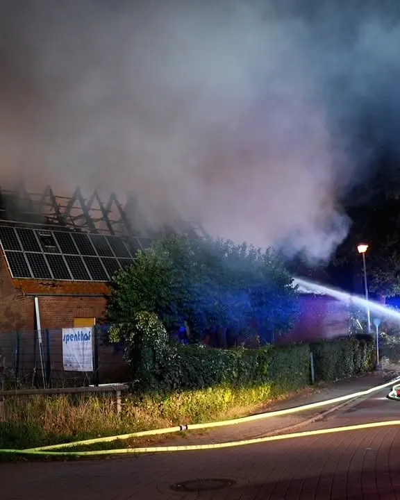 Eine Feuerwehr löscht ein Gebäude, aus dessen Dach dichter Rauch aufsteigt. Ein Wasserschlauch spritzt auf die Flammen.