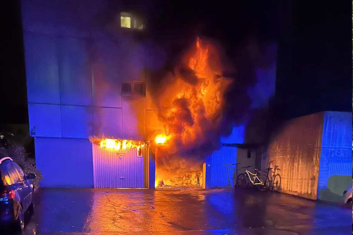 Flammen und dichter Rauch steigen aus einem Garagentor, während das umliegende Gebiet in der Nacht dunkel ist. Mehrere Fahrräder sind links daneben sichtbar.