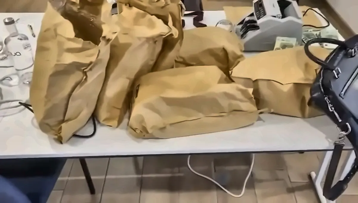 Auf einem Tisch befinden sich mehrere in braunes Papier eingewickelte Pakete neben einer Geldzählmaschine und Bargeld.